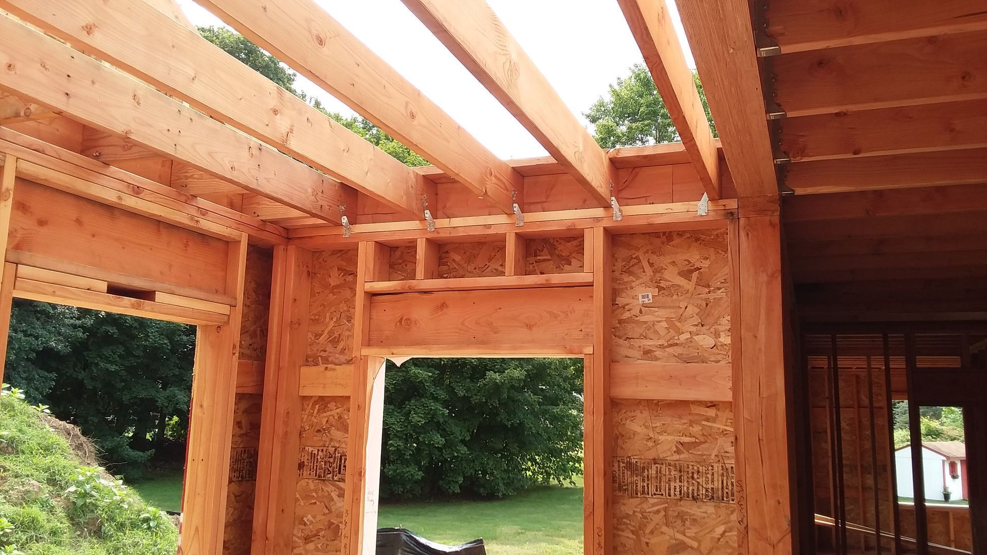 Wooden house frame under construction, featuring exposed beams, plywood walls, and door openings, outdoors on a sunny day.