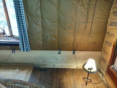Room interior with exposed insulation, wooden paneling, and a lamp. A window with a curtain is on the left.