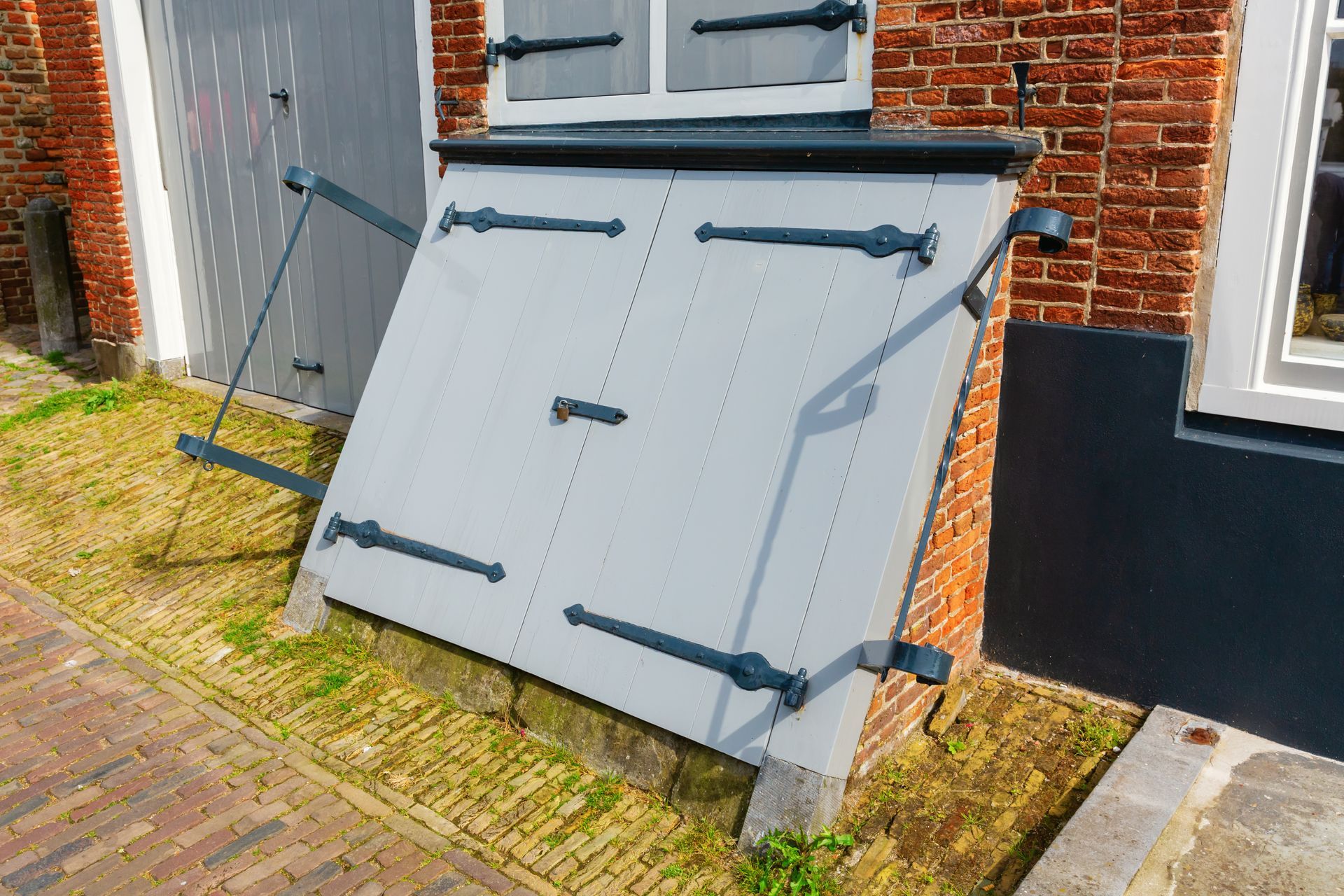 Gray cellar doors with black hardware, next to brick wall and white window.