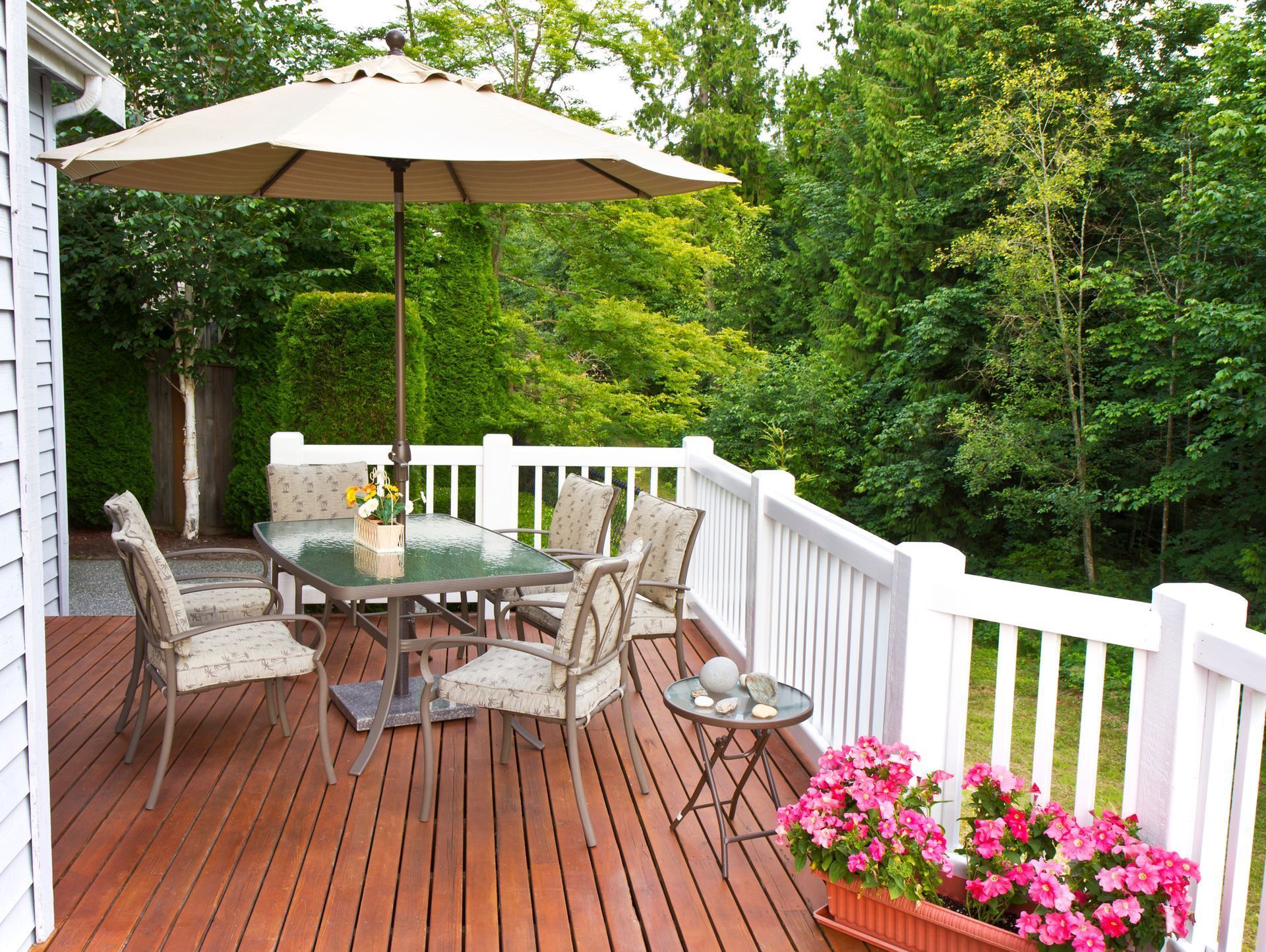 Wooden deck with outdoor furniture under a beige umbrella, surrounded by greenery.