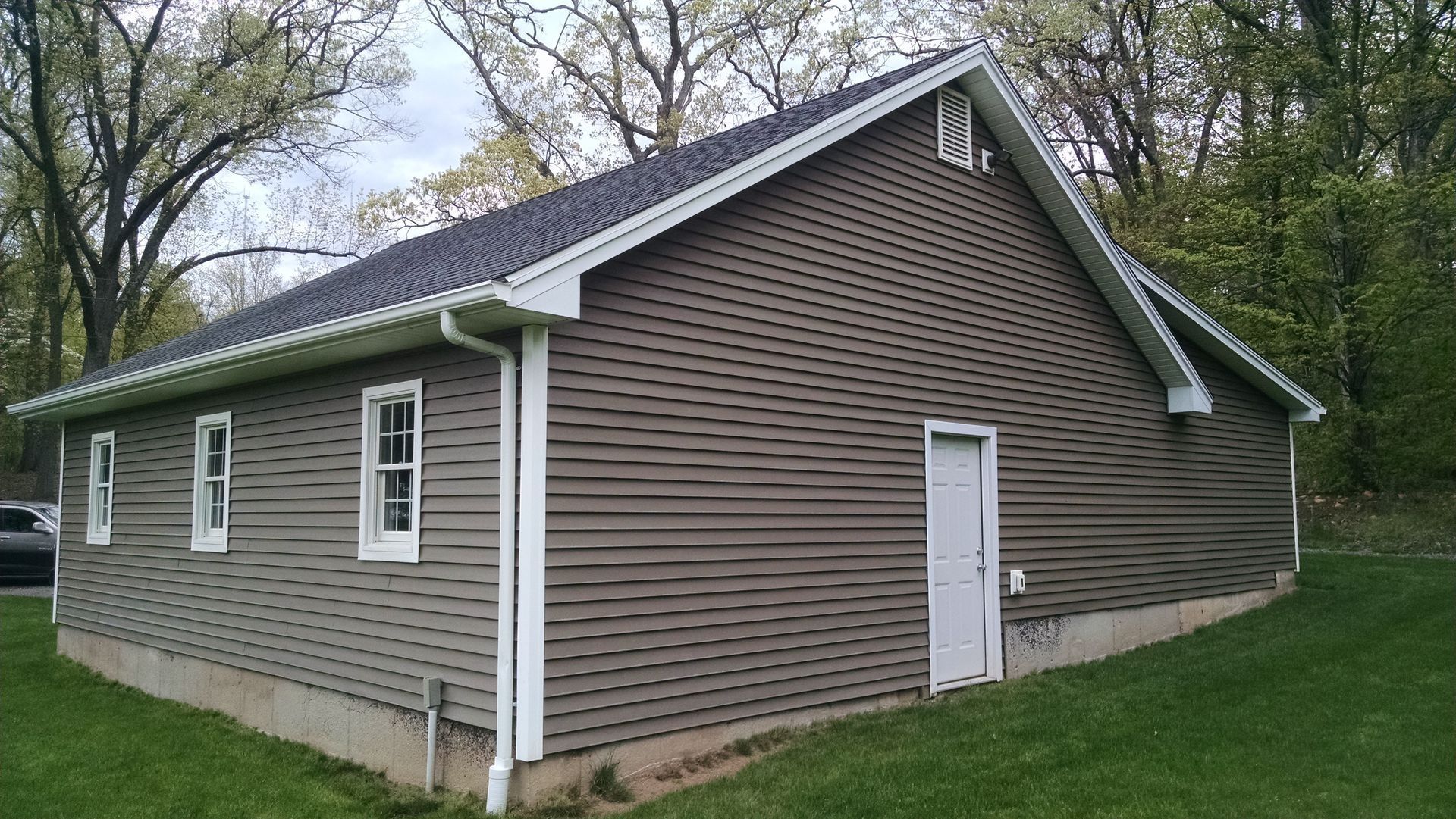 Brown-sided building with white trim, windows, and door. Set on a grassy area, trees in background.