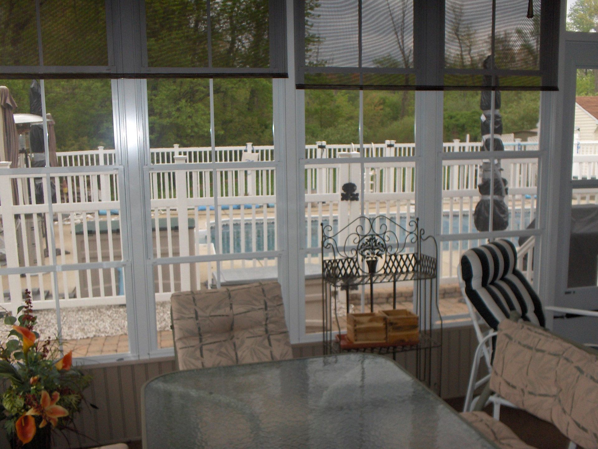 Sunroom interior with view of a pool, white fence, and greenery. Furniture is visible.