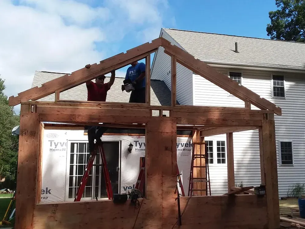 Construction workers building a wooden structure attached to a house; blue sky background.