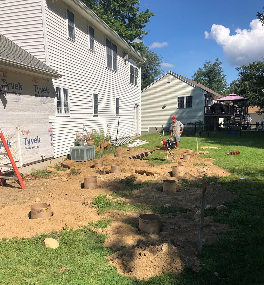 Construction site with house. Holes dug in dirt. Person operating equipment. Sunny day.