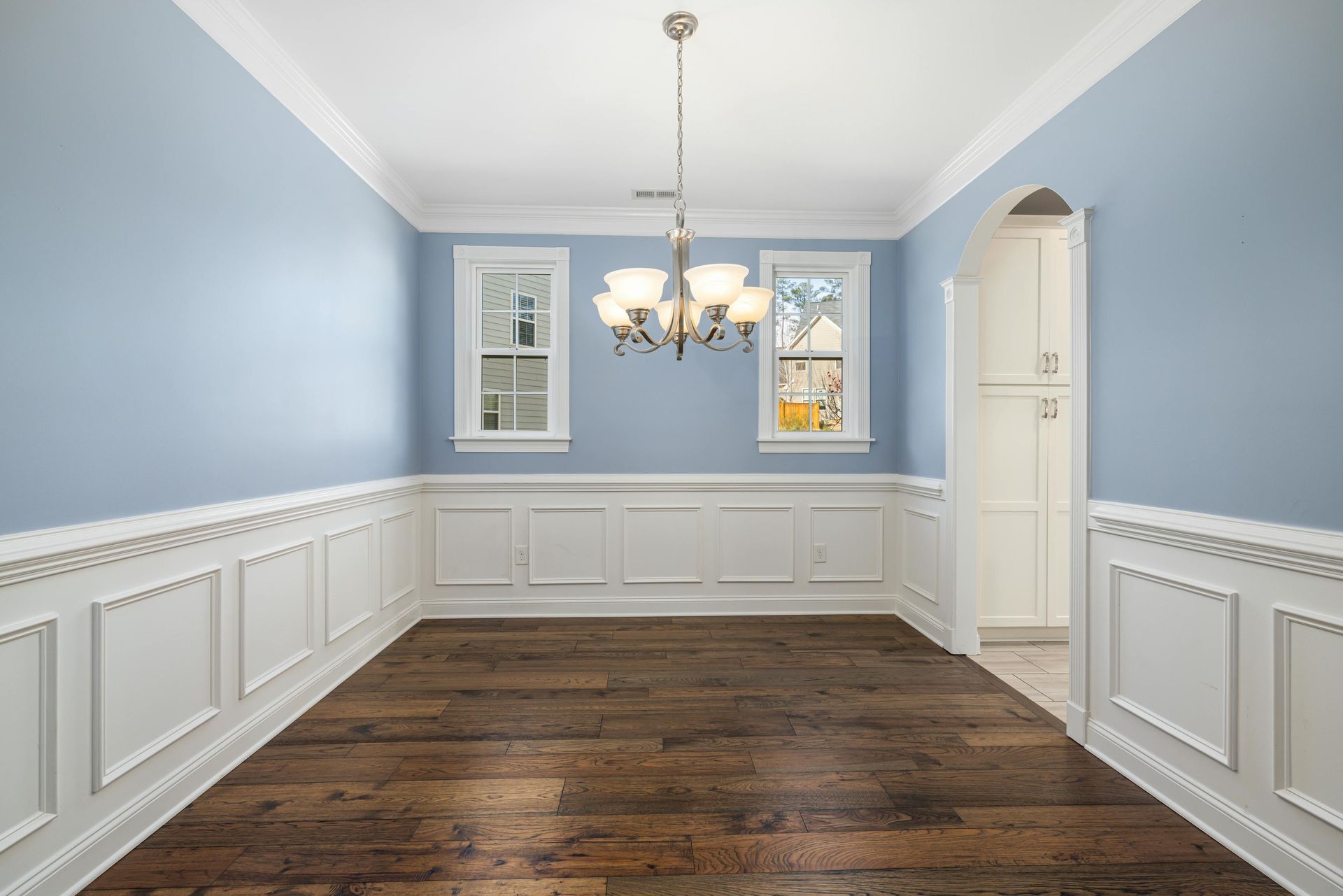 Empty dining room with blue walls, white trim, hardwood floor, chandelier, and arched doorway.