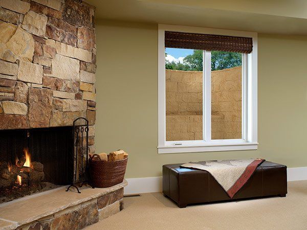 Fireplace and window in room; stone facade, tan walls, brown woven shade, and brown leather ottoman.