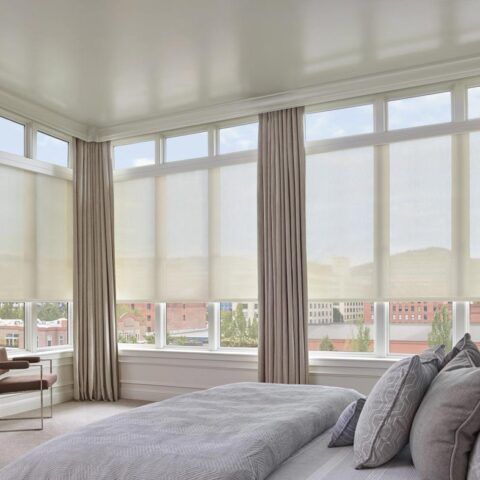 Bright bedroom with large windows, sheer curtains, and a bed with gray bedding overlooking a city view