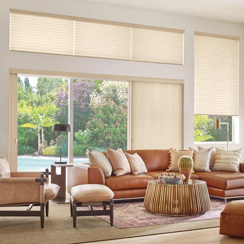 Modern living room with tan sectional, woven coffee table, and large windows with blinds overlooking greenery