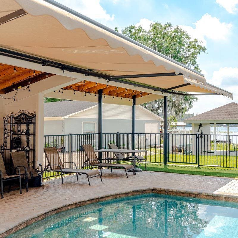 Covered patio with lounge chairs beside a backyard pool and lake view