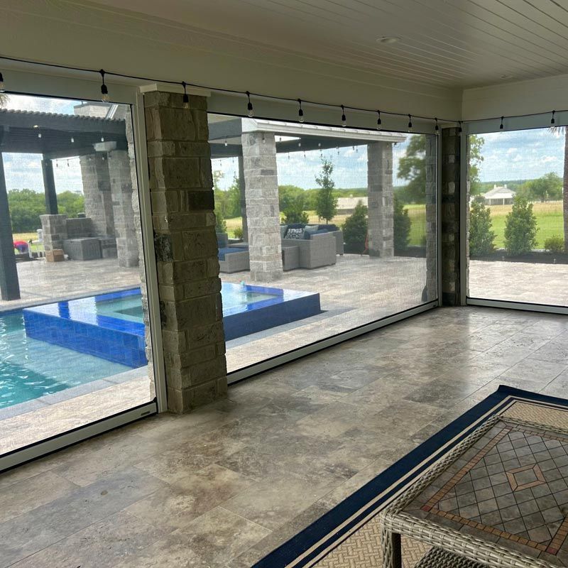 Covered patio with floor-to-ceiling windows overlooking a pool and stone patio seating area