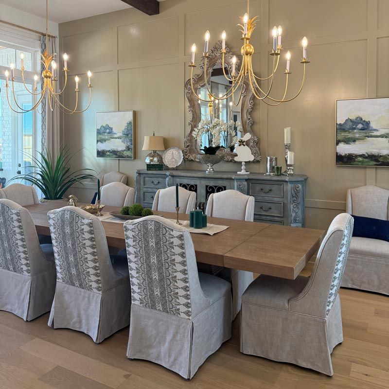 Formal dining room with a long wood table, gray slipcovered chairs, chandeliers, and a sideboard with decor.
