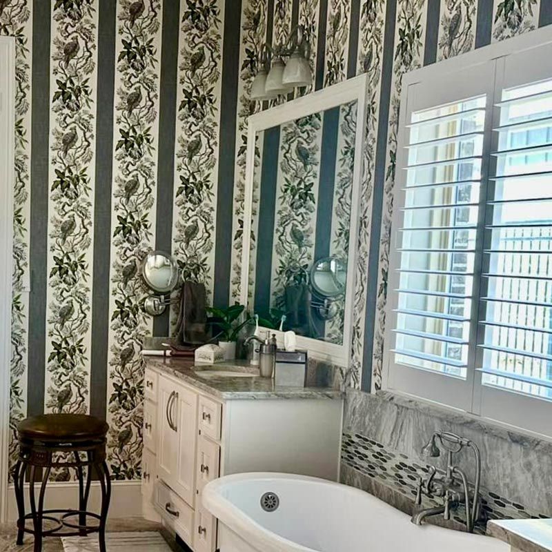 Elegant vintage bathroom with floral wallpaper, white shutters, marble vanity, and freestanding tub