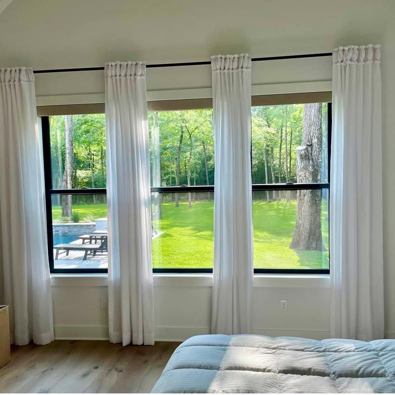 Bright bedroom with white curtains, large windows, and a green wooded view outside
