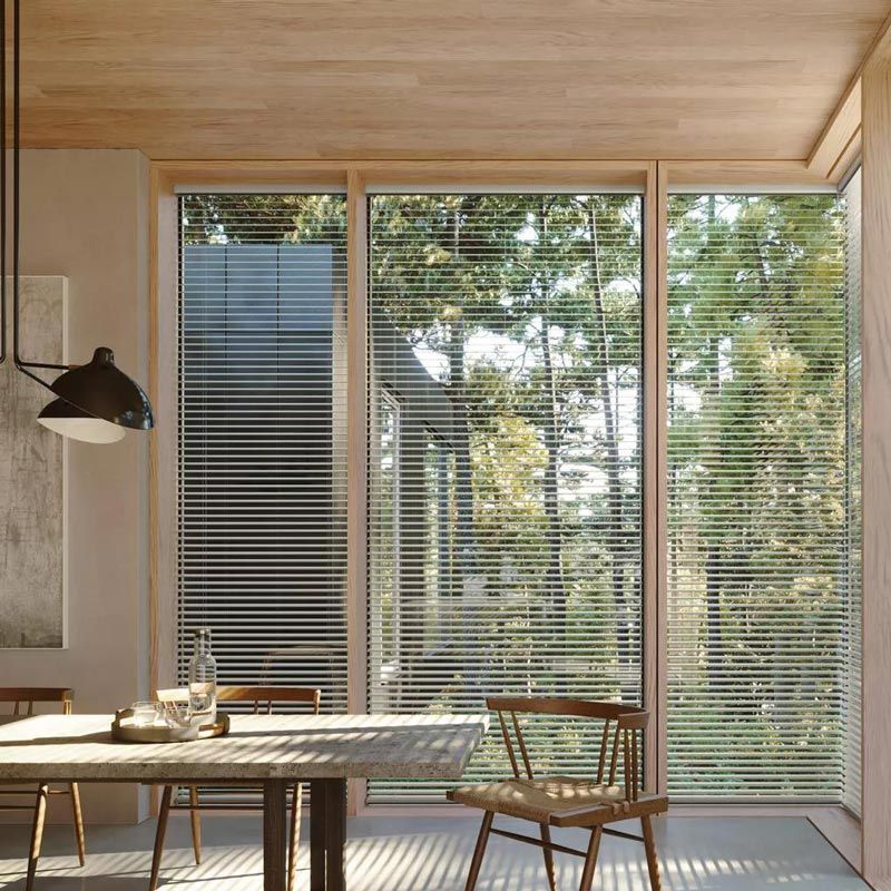 Sunlit modern dining room with wooden table, chairs, and large glass doors with blinds overlooking greenery