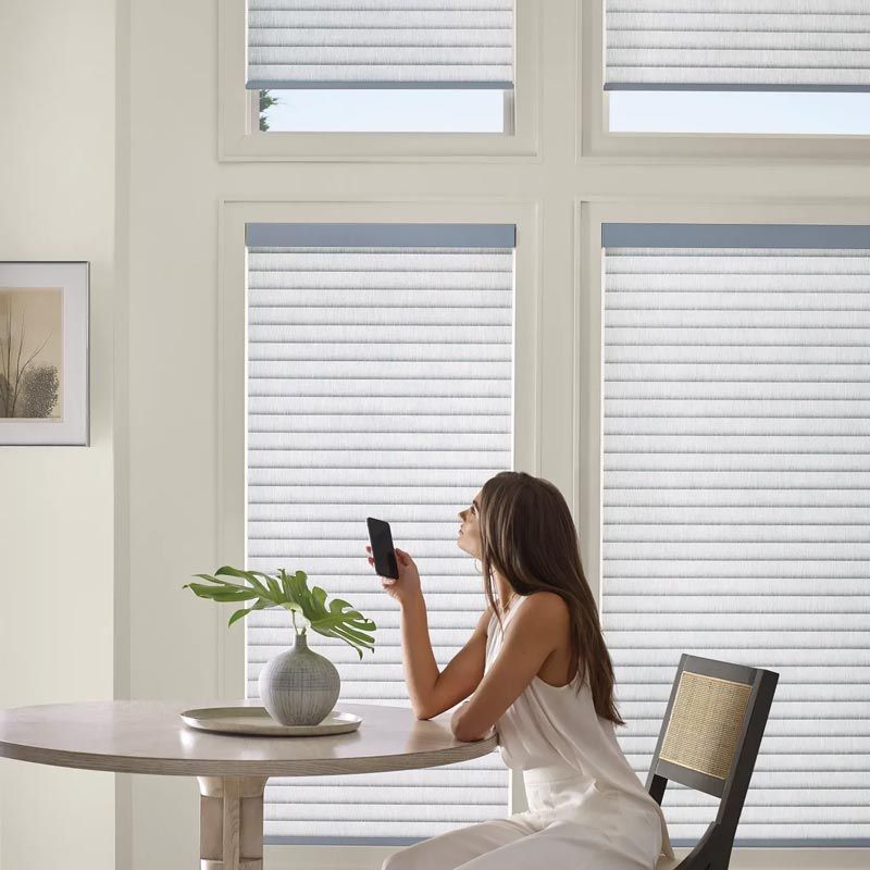 Woman sitting at a table by large windows, looking at her phone in a bright room.