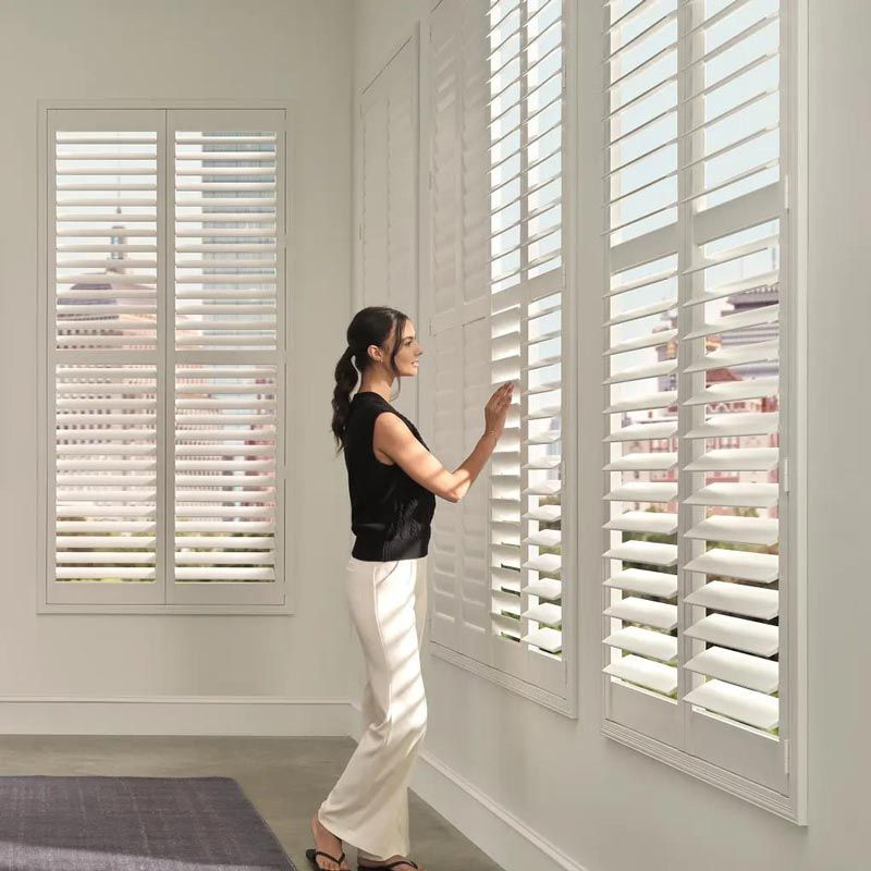 Woman adjusting white window shutters in a bright room