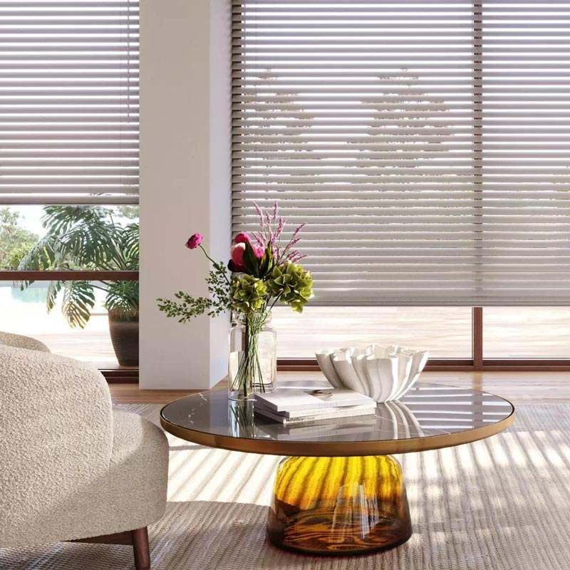 Modern living room with round glass coffee table, vase of flowers, bowl, and sunlight through blinds
