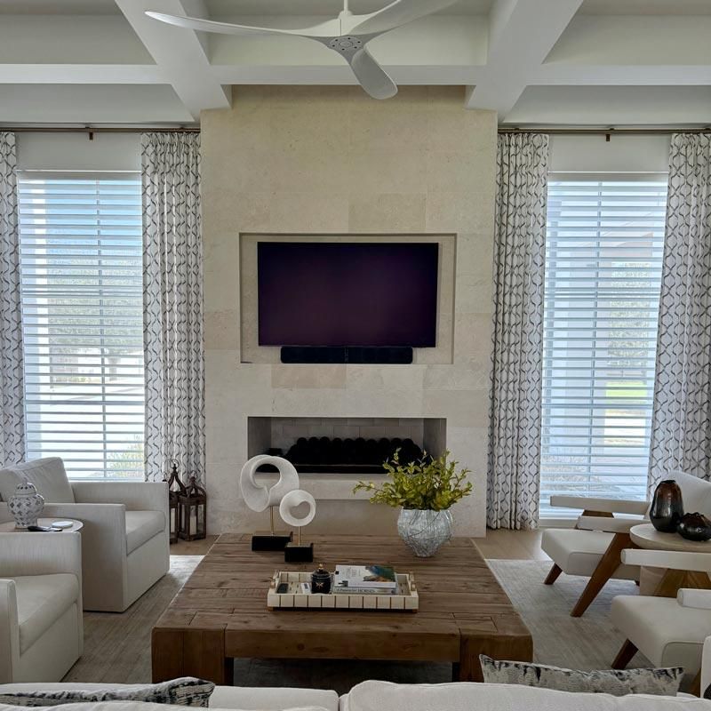 Bright living room with white sofas, wood coffee table, and TV over a fireplace between tall windows.