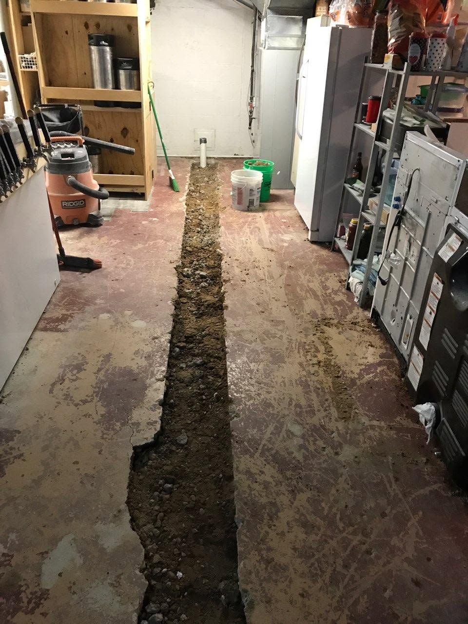 Narrow trench cut through a basement floor, with tools and materials around it.