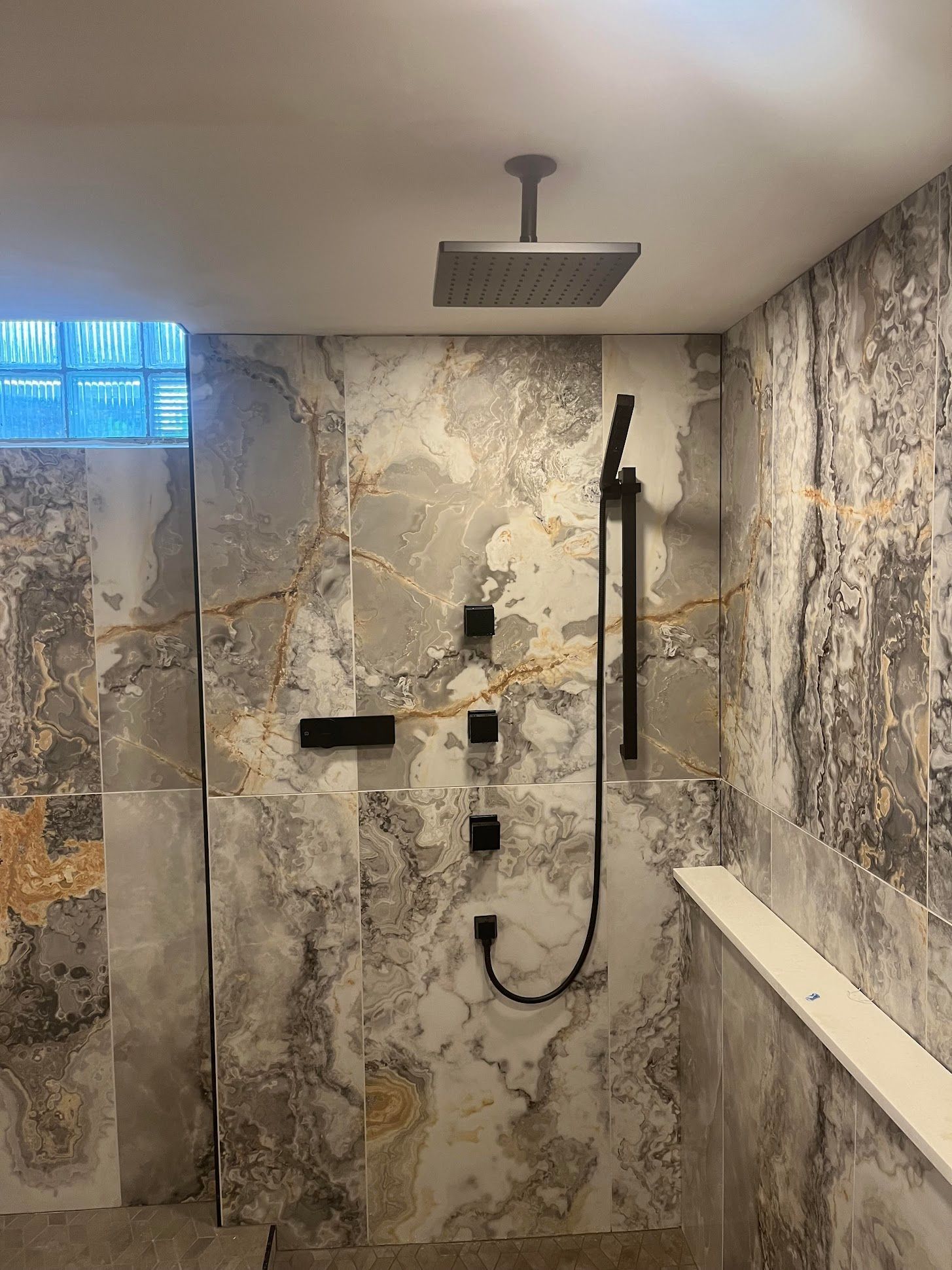 Modern marble-tiled shower with rainfall head, handheld sprayer, and built-in shelf.