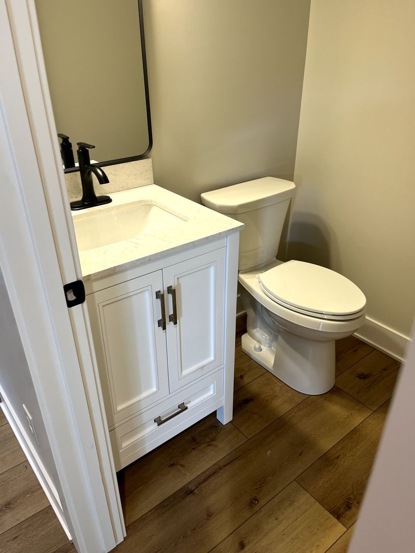 Small bathroom with white vanity, toilet, mirror, and wood-look floor.
