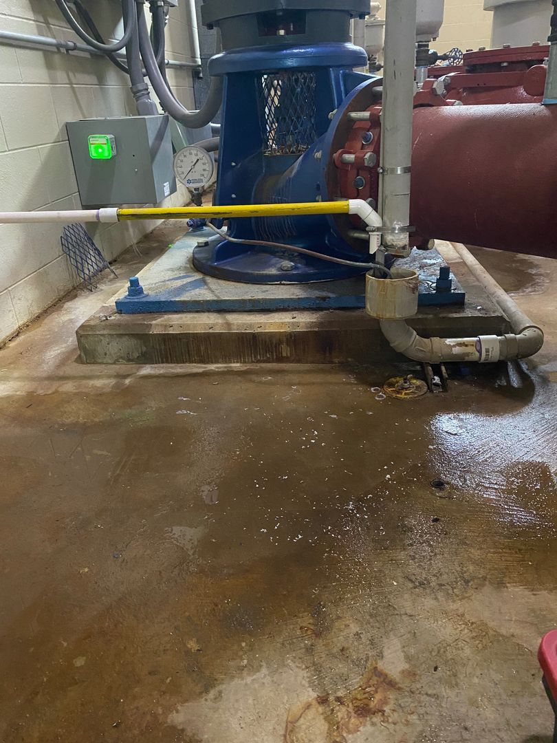 Industrial utility room with blue machinery, pipes, and a wet concrete floor behind a yellow barrier line