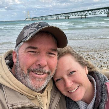 John & Penny Schnittker selfie on a rocky beach with the Machinaw Bridge in the background.