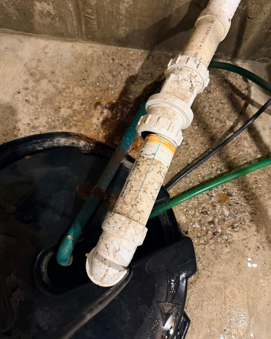 Dirty white PVC pipe fittings and green hoses beside a wet black basin on a concrete floor