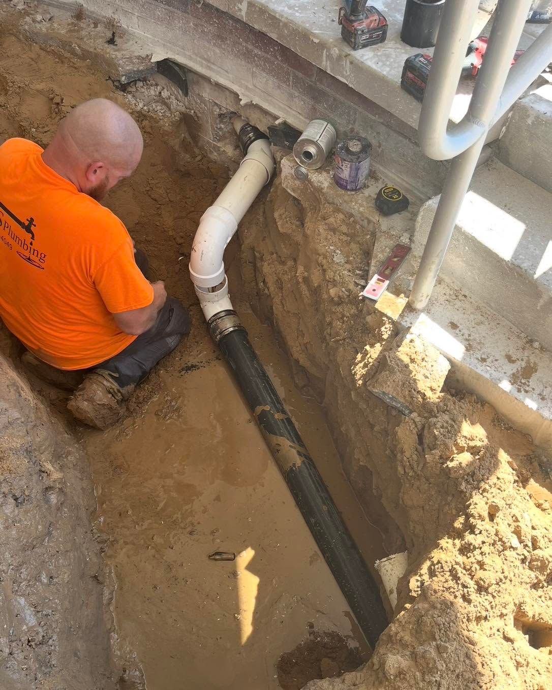 Worker in orange shirt kneels in a trench, installing underground piping near a wall.