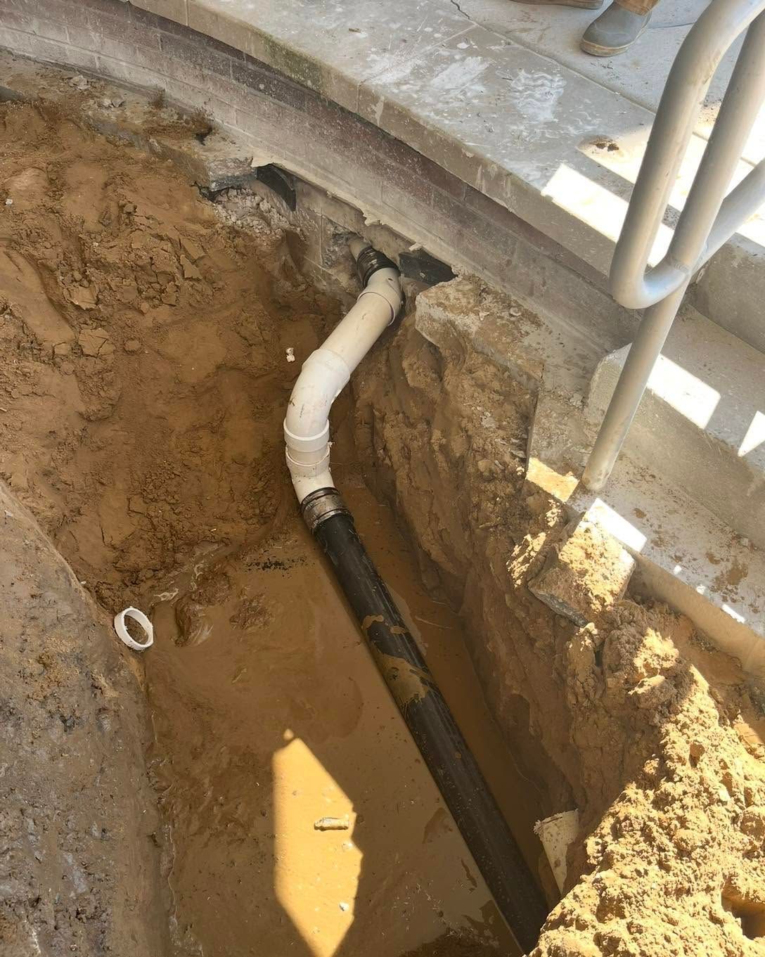 Exposed black pipe with white elbow in a muddy trench beside a concrete slab and metal railing