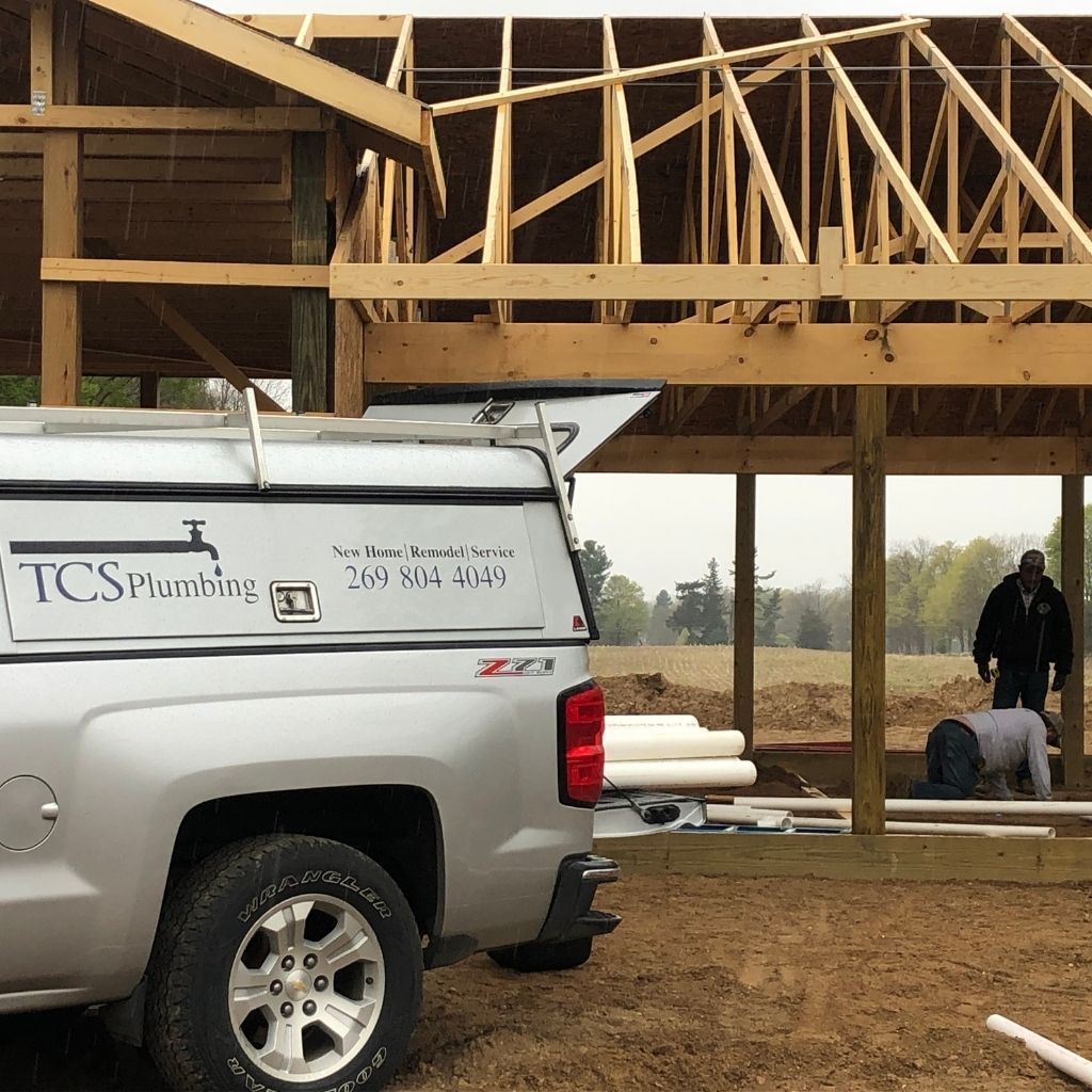 Silver plumbing truck at a house construction site with wood framing and workers in the background
