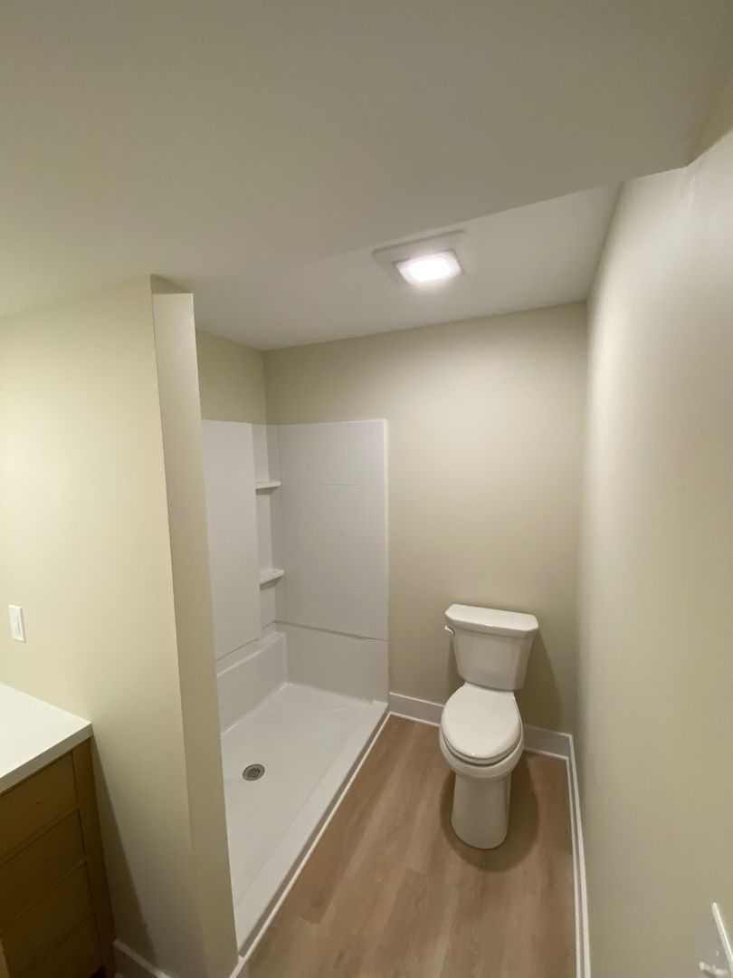 Small beige bathroom with toilet, shower stall, and wood-look floor under bright ceiling light