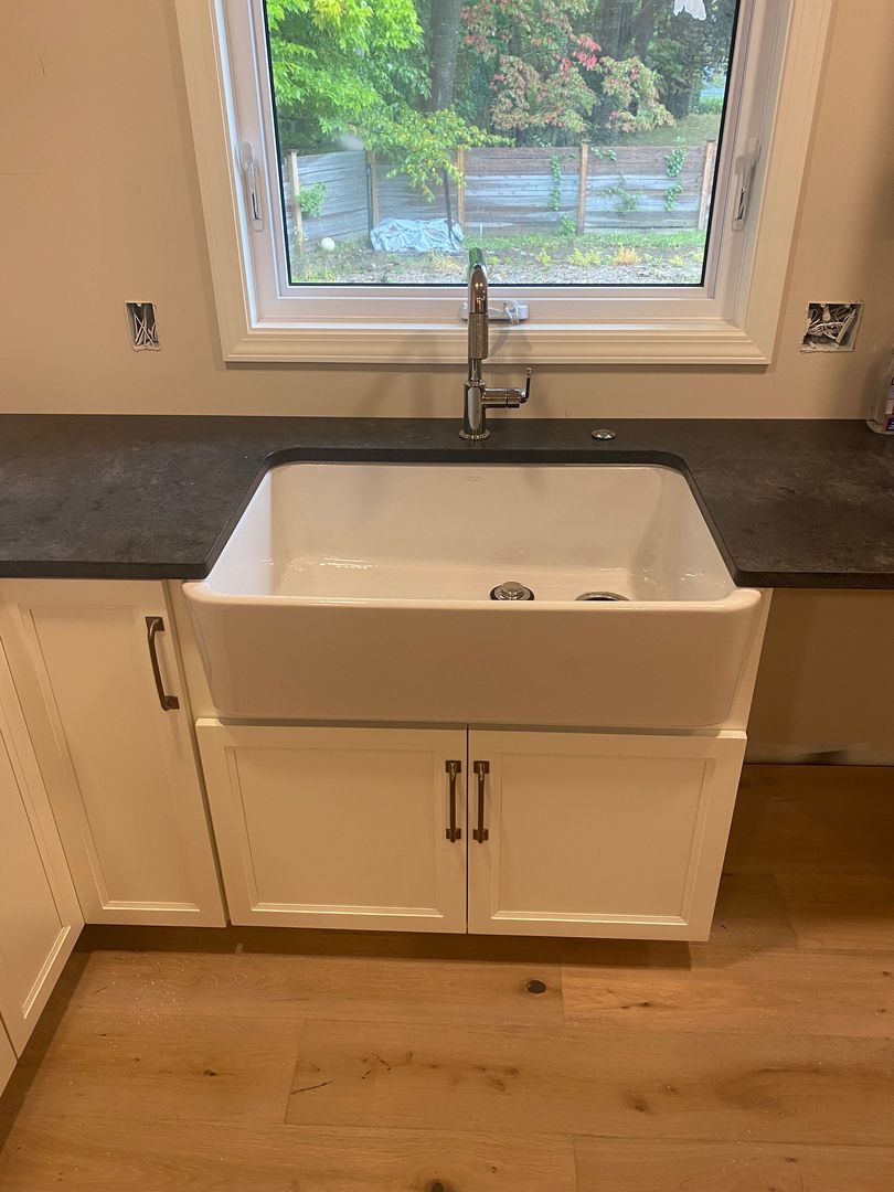 Kitchen sink under a window with dark countertops, white cabinets, and wood flooring.