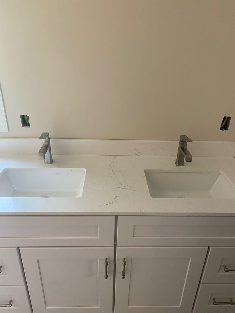 Double vanity bathroom sink with white countertop and cabinets, two silver faucets, and unfinished wall outlets