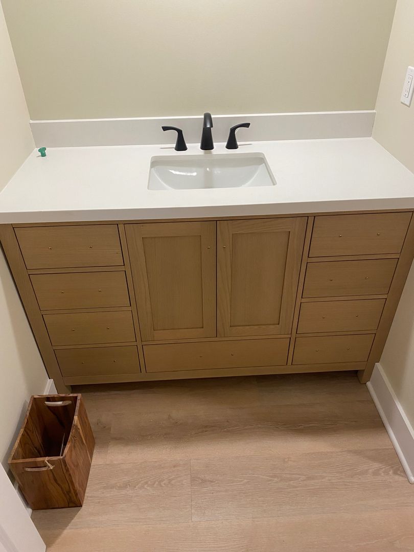 Bathroom vanity with white countertop, sink, black faucet, and wooden cabinet doors against beige walls.