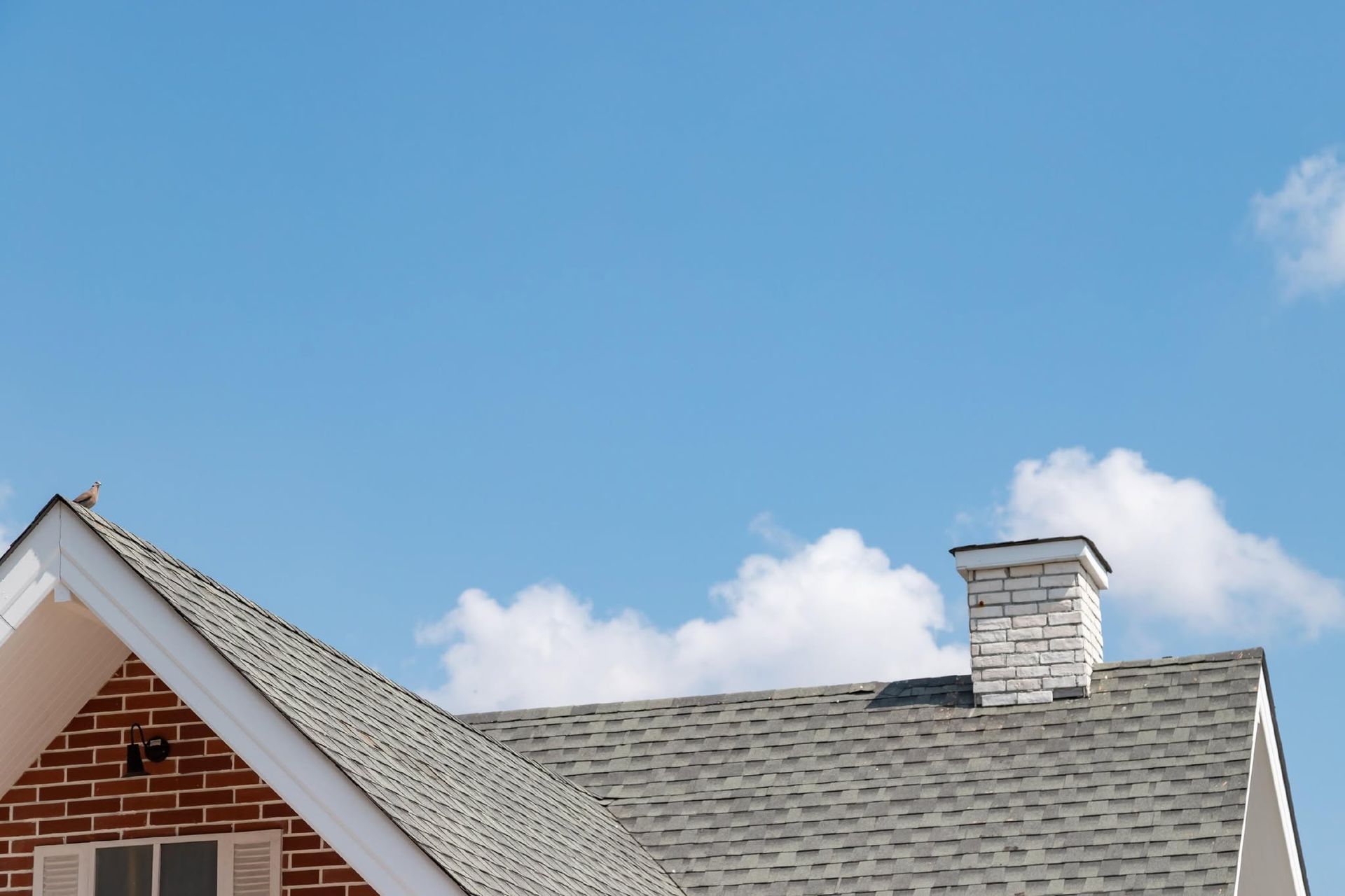 Roofline with gray shingles and a white chimney against a blue sky with clouds