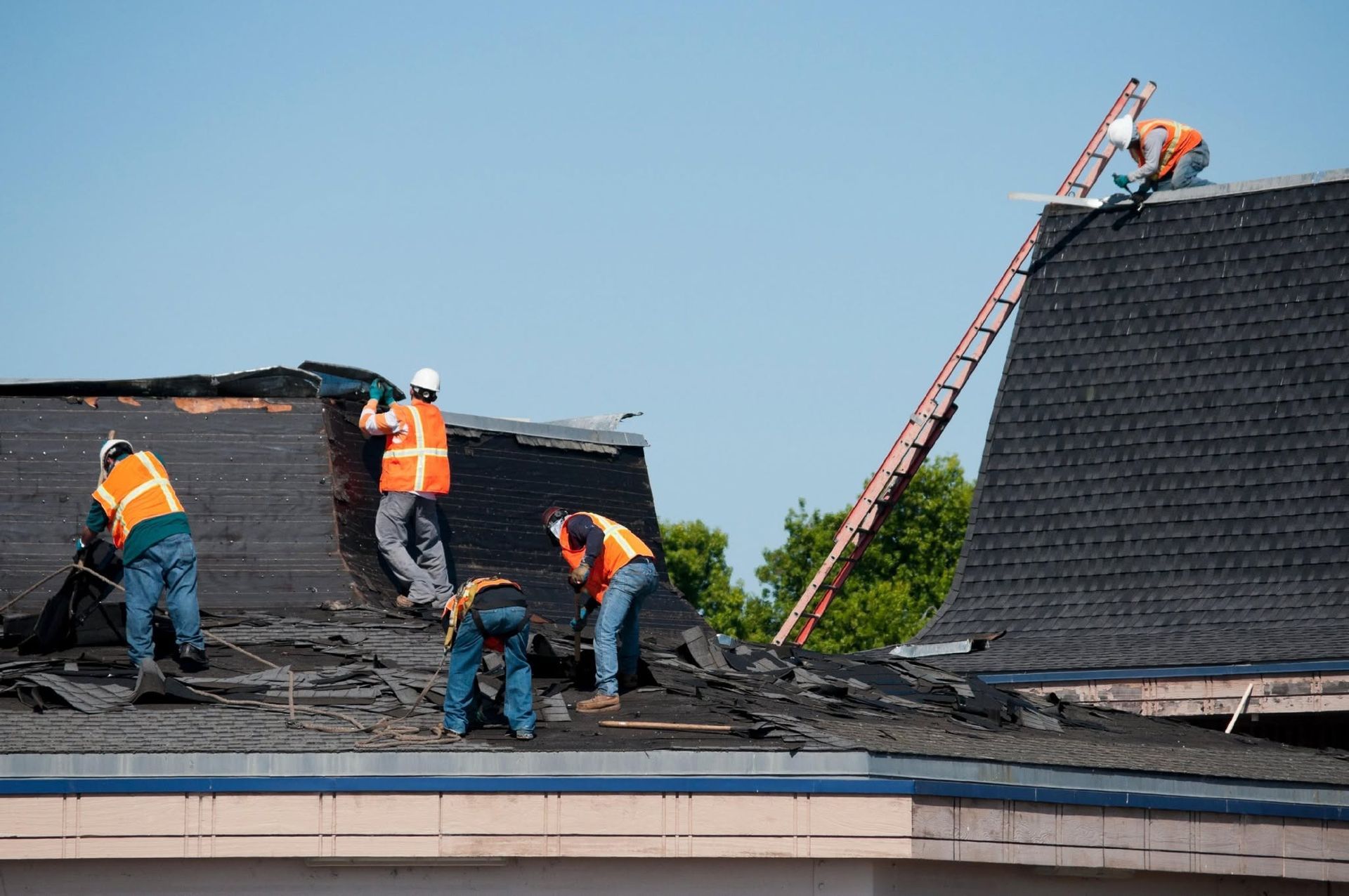 Roofing workers repairing a flat roof, with one climbing a ladder on a sunny day.