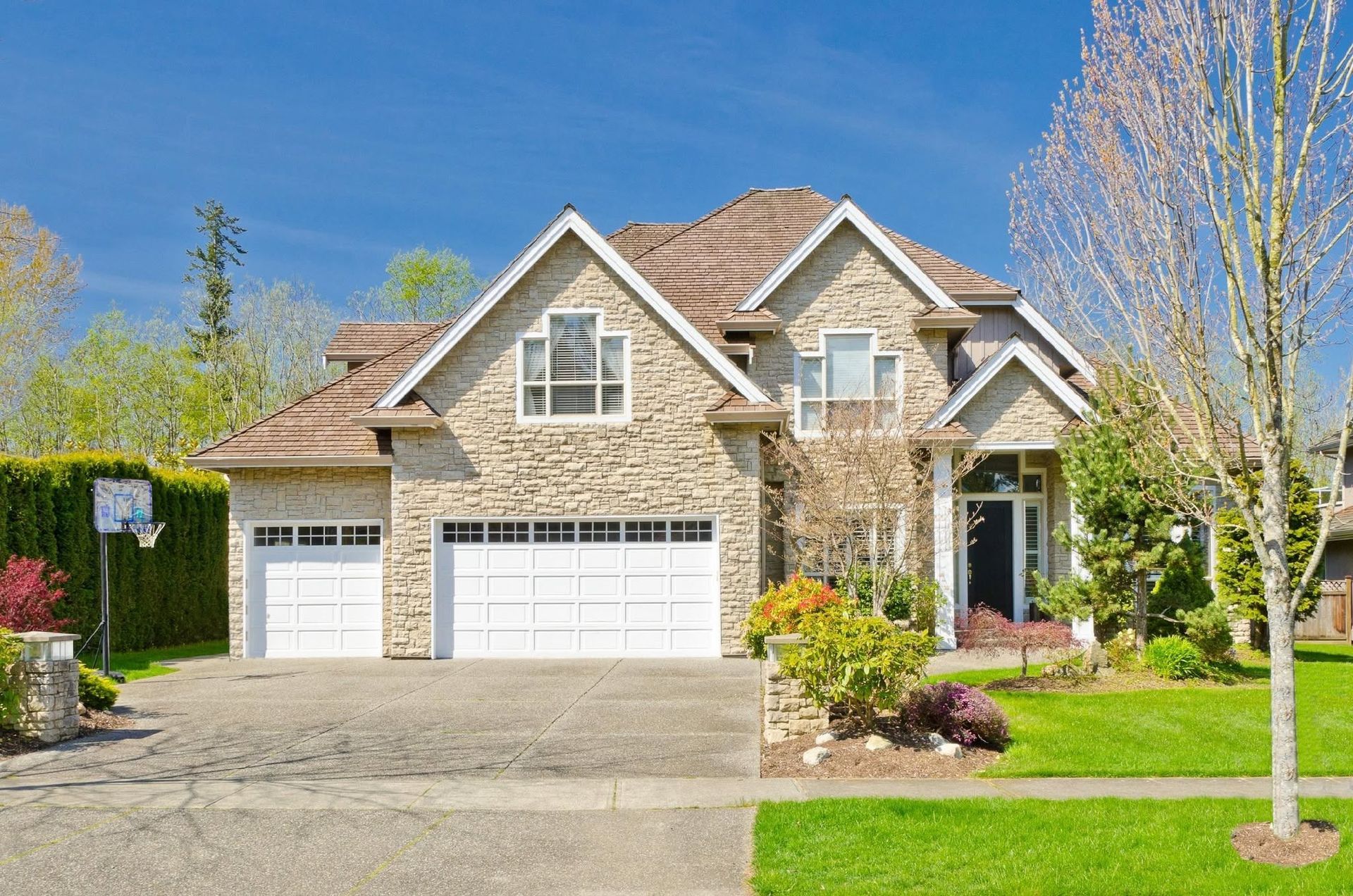 Suburban stone house with three-car garage, driveway, and manicured lawn under a clear blue sky