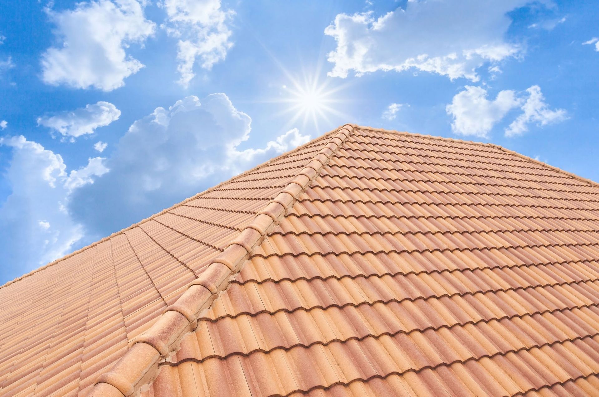 Terracotta roof tiles against a bright blue sky with the sun shining above the peak