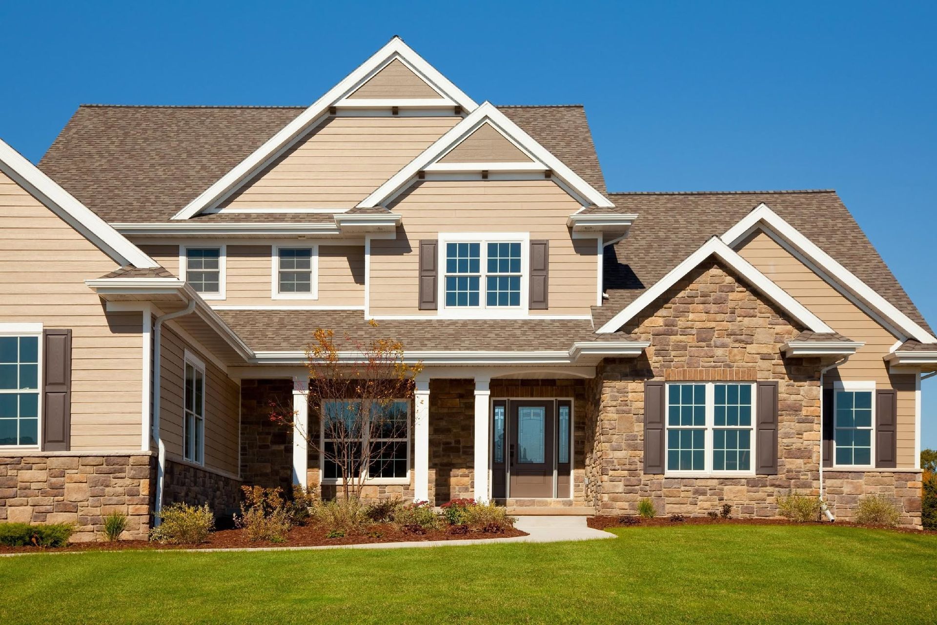 Large beige suburban house with a brown roof, white trim, front porch, and green lawn under a blue sky
