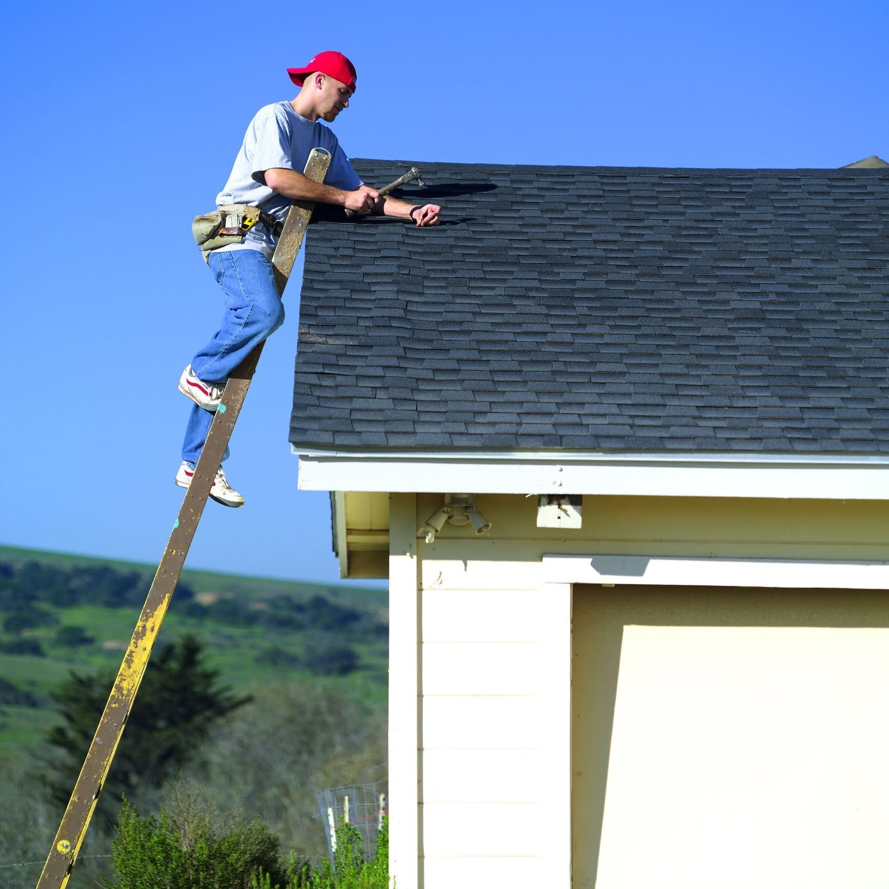 Worker on a roof shingling the edge of a house, standing on a ladder under a blue sky.
