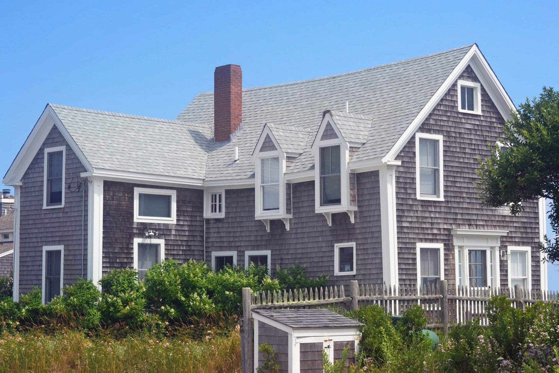 Gray shingled coastal house with white trim, dormer windows, and a red chimney behind a white picket fence