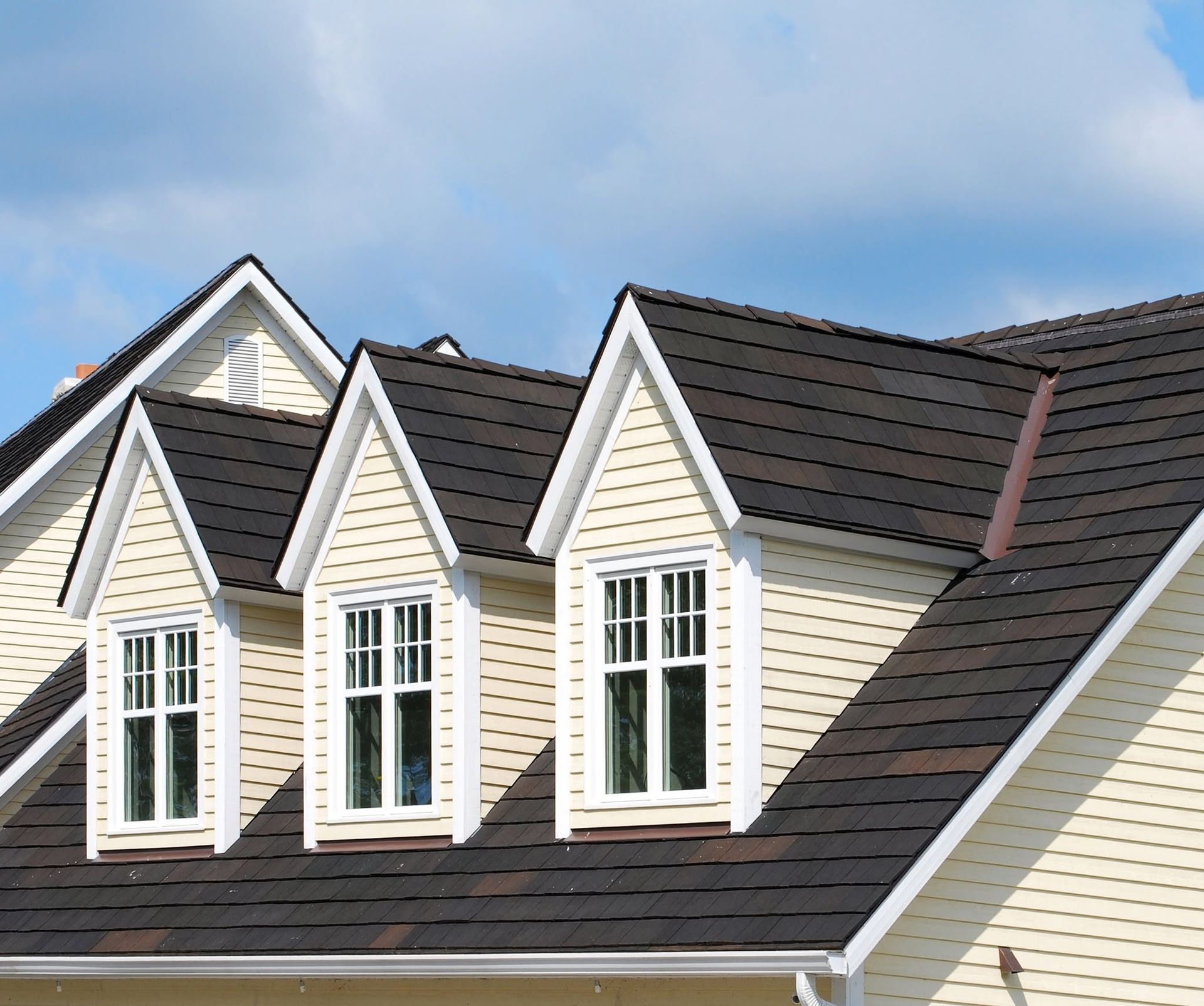 Row of cream dormer windows with white trim on a dark shingled roof against a blue sky