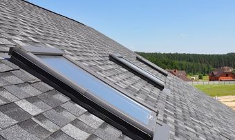 Roof with two black skylights on gray shingles under a clear blue sky