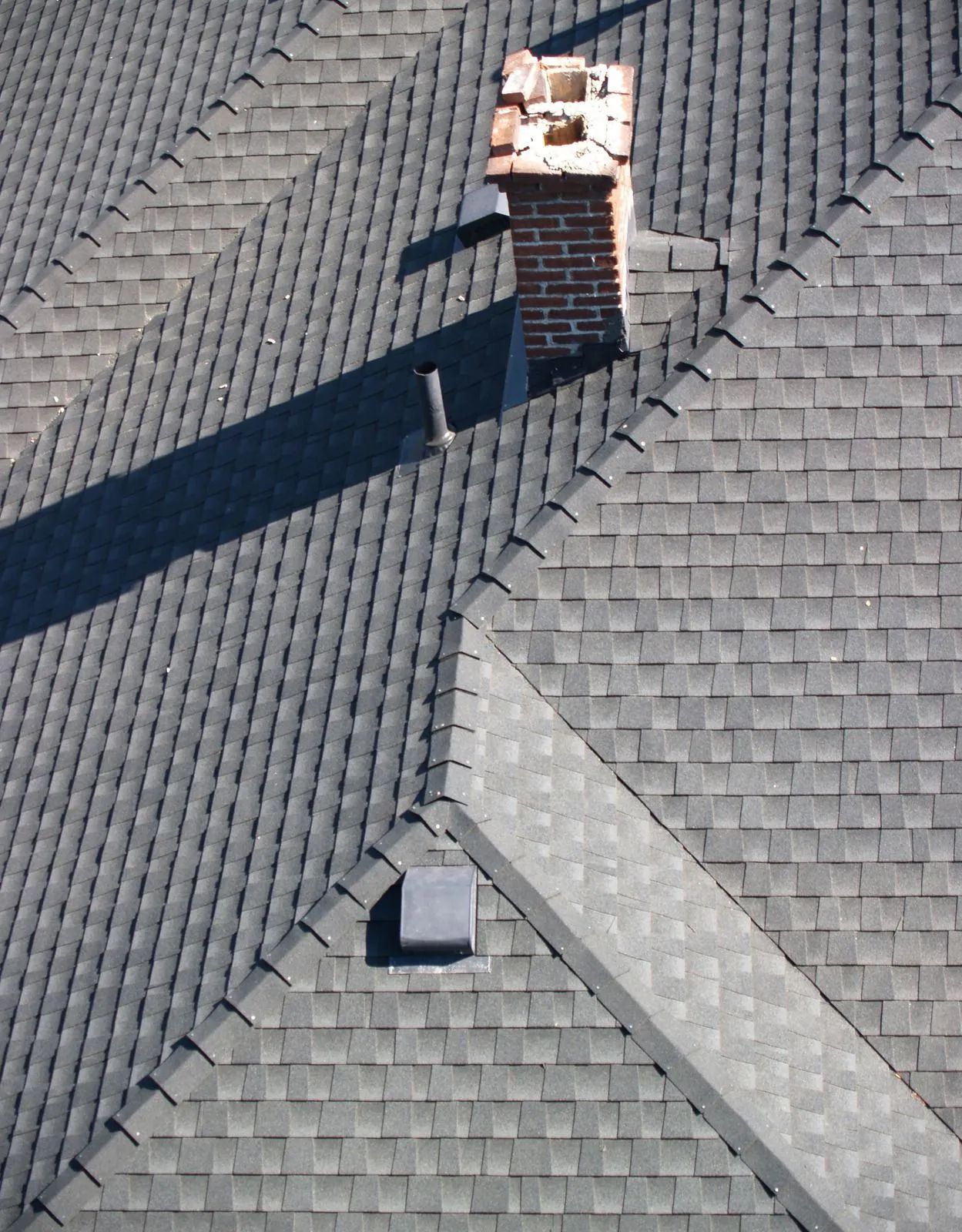 Gray shingled roof with a brick chimney and small skylight under bright sunlight