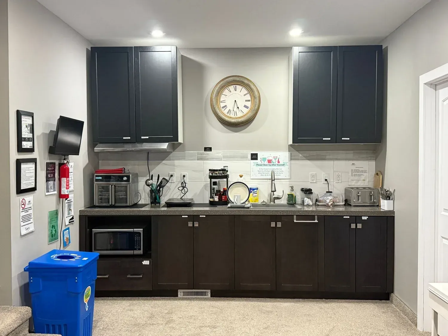 Kitchen with dark cabinets, clock above sink, countertop appliances, and a blue recycling bin on the left.