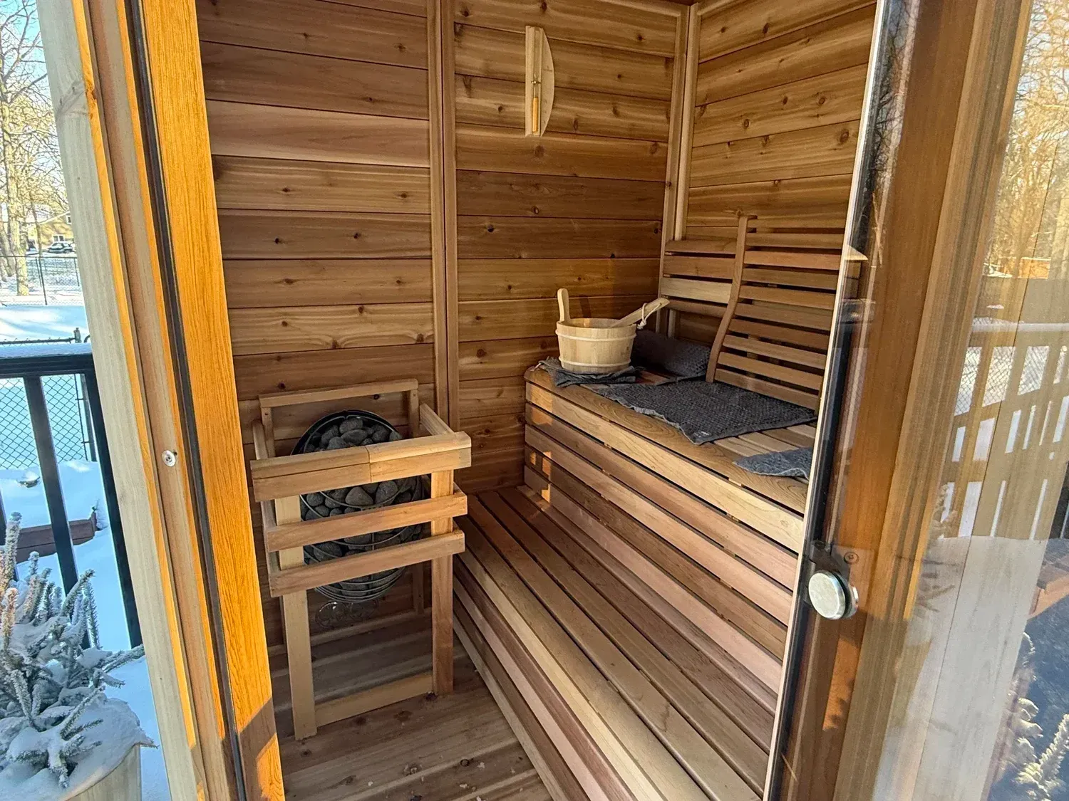 Wooden sauna interior with bench seating, bucket, and snow visible outside the doorway