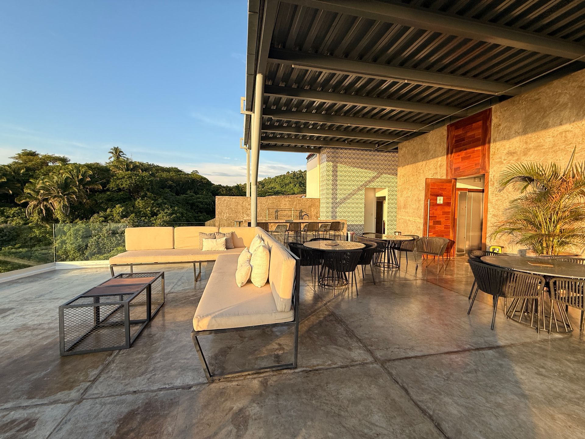 Oceanfront terrace with black tables and chairs under a metal roof, blue sky and sea beyond.
