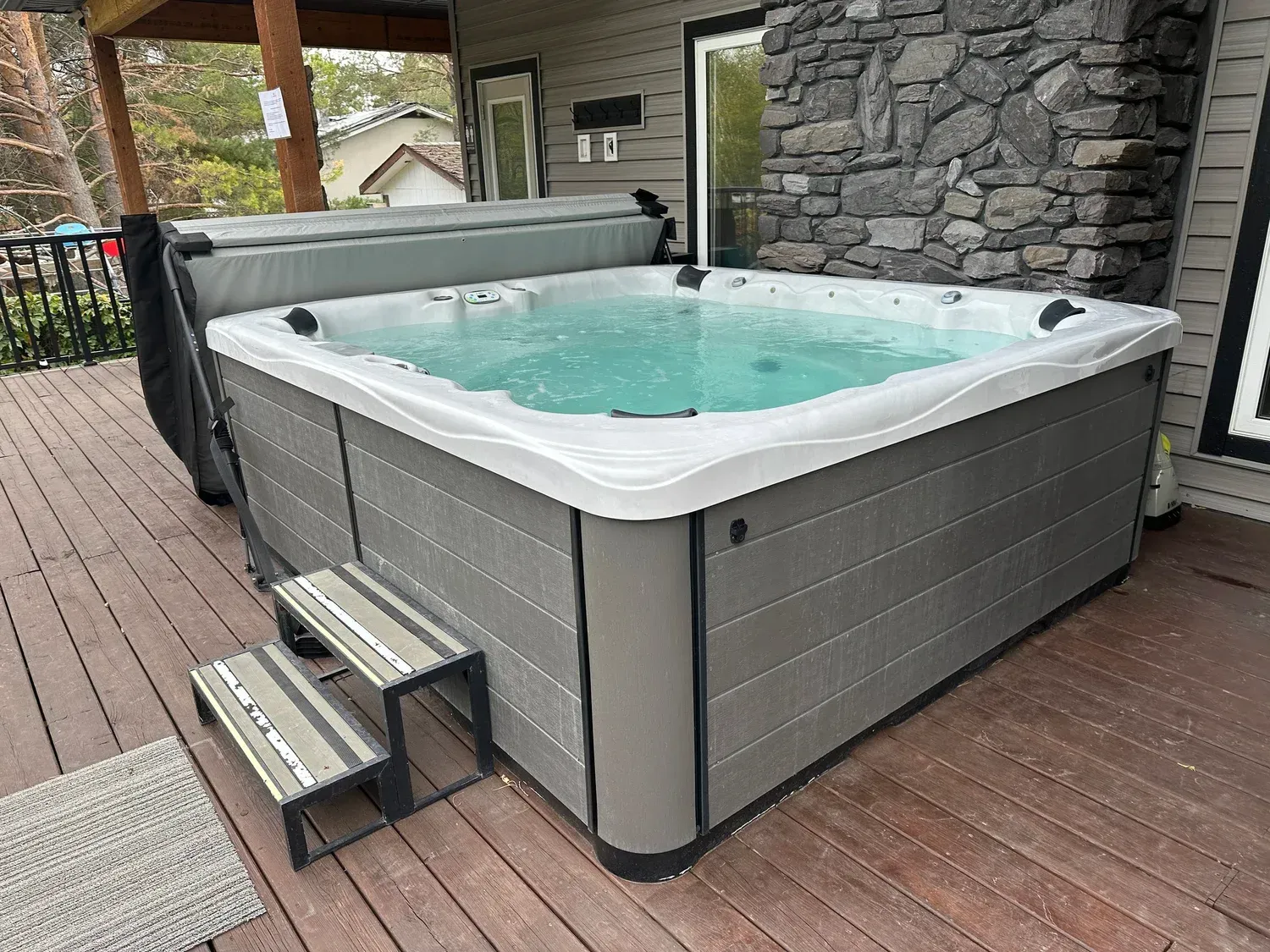 Gray hot tub on a wooden deck beside a stone wall, with steps and turquoise water.