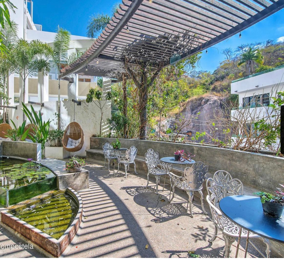 Sunlit patio with metal café tables, striped pergola shadows, and a small pond beside lush greenery